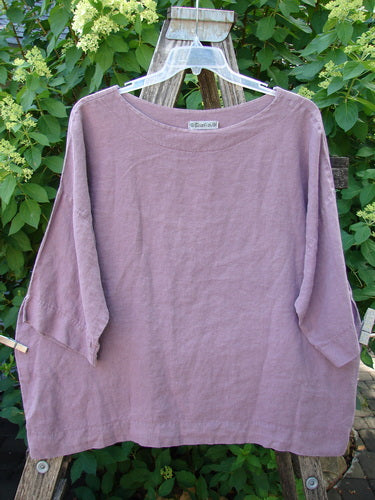 Barclay Linen Lantern Boxy Unpainted Dark Mauve Size 1 top with a wide boat neckline and three-quarter sleeves, draped on a wooden ladder amidst lush foliage, showcasing its boxy, slightly cropped design.