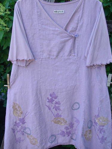 Barclay Linen Lace Blooming Tunic Dress in Leaf Vine Lavender, featuring a V-neck with a button, ruffled sleeves, flared skirt, and leaf motifs, hanging outdoors against green foliage.