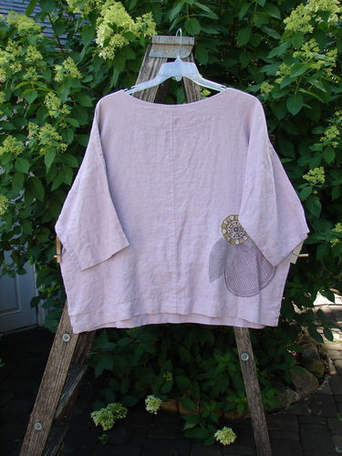 Barclay Linen Lantern Boxy Giant Blossom Hydrangea Size 1: A pale-pink, loose-fitting linen top with 3/4 sleeves and floral appliqués, displayed on a wooden ladder amidst lush greenery and hydrangeas.