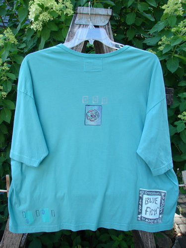 Barclay Short Sleeved Crop Box Tee Center Rose Aqua Size 3, featuring an airy, oversized square shape, ribbed neckline, wider sleeves, and Blue Fish stamp, hanging against a green shrub backdrop.
