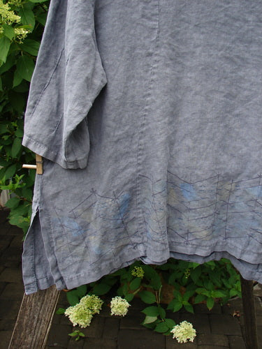 Barclay Linen Pinch Sleeve Curve Tie Back Top Border Twig Grey Size 1, displayed on a wooden rail, showcases a blue hand-drawn triangular pattern at the hem against lush green foliage.