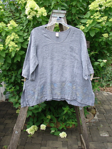 The Barclay Linen Pinch Sleeve Curve Tie Back Top Border Twig Grey Size 1 elegantly drapes over a wooden ladder, showcasing its loose fit and lightweight texture amidst a serene garden backdrop.