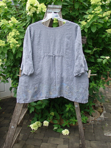 Barclay Linen Pinch Sleeve Curve Tie Back Top Border Twig Grey Size 1, a loose-cut, long-sleeved blouse, hangs on a wooden ladder, showcasing its simple round neckline and flared hem.