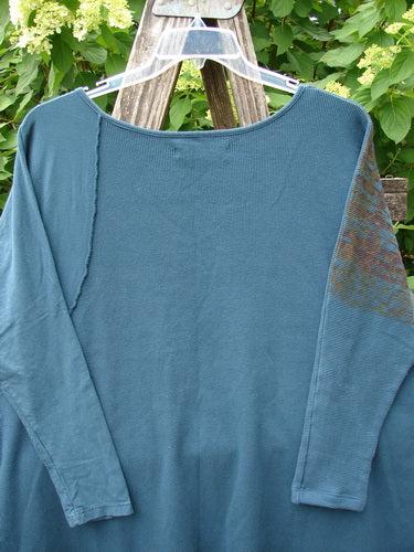 Barclay Thermal Monte Verde Vented Tunic Grid Blend Tealen OSFA hangs on a ladder, showcasing its rounded neckline, slim fitted sleeves, and unique exterior stitchery against a lush, floral backdrop.