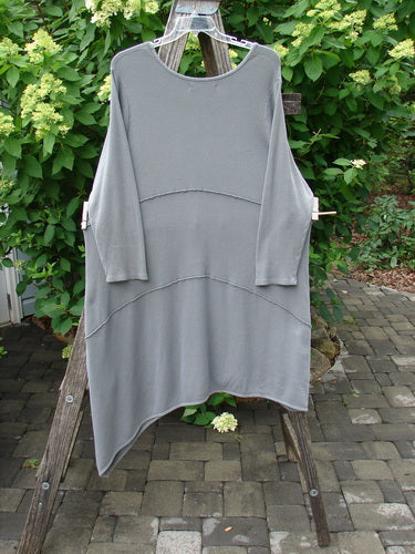 Barclay Thermal Exterior Stitch Three Tier Dress, Forest Friend Grey Day Size 2, hangs on a wooden A-frame. This loose-fitting dress features an asymmetrical hem and visible pulls, set against a lush garden backdrop.