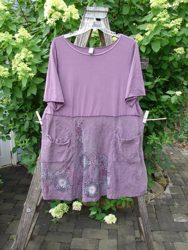 Barclay Linen Cotton Two Pocket Tie Back Square Dress Bloom Burst Plum Size 1, featuring a loose fit, short sleeves, front patch pockets, and a subtle floral print, displayed on a wooden stand outdoors.