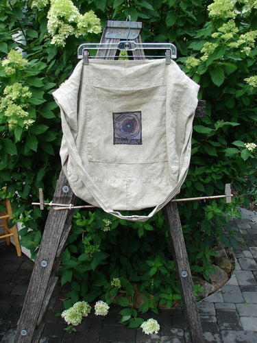 Barclay Patched Linen Pocket Tote Sling in Wheat displayed on a wooden trestle, featuring a wide shoulder strap, spiral graphic, and signature patch, set against lush greenery and white hydrangeas.