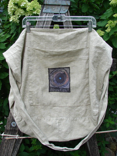 Barclay Patched Linen Pocket Tote Sling Window Door Wheat OSFA: A light-tan linen bag with a Bluefish patch, large front pocket, and long strap, displayed on a ladder against green foliage.