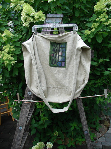 Barclay Patched Linen Pocket Tote Sling Window Door Wheat OSFA draped over a rustic wooden ladder, featuring abstract geometric patchwork in greens, blues, and purples, set against lush outdoor foliage.