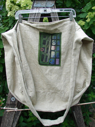 Barclay Patched Linen Pocket Tote Sling Window Door Wheat OSFA, hanging from a wooden A-frame, showcases a colorful grid-style patch pocket. Features a slouchy design with a wide shoulder sling.