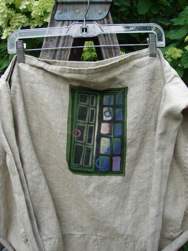 Barclay Patched Linen Pocket Tote Sling Window Door Wheat OSFA: A light-tan hoodie with abstract panel graphic, featuring a dark-green bordered design with colorful shapes, hung on a wooden support outdoors.