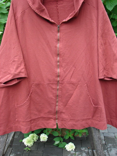 Barclay PMU Patched Hooded Postcard Zipper Jacket Vest in Deep Brick, featuring a full front zipper, kangaroo-style pocket, three-quarter sleeves, and a graphic rear patch, hanging amidst white hydrangeas.