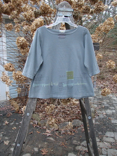 Muted-green 2000 Parklands Tee Top Latitude Iron Size 1 with three-quarter sleeves and a slightly flared shape, displayed on a hanger against a rustic background, featuring neat X-stitched patches and a pale-green graphic.
