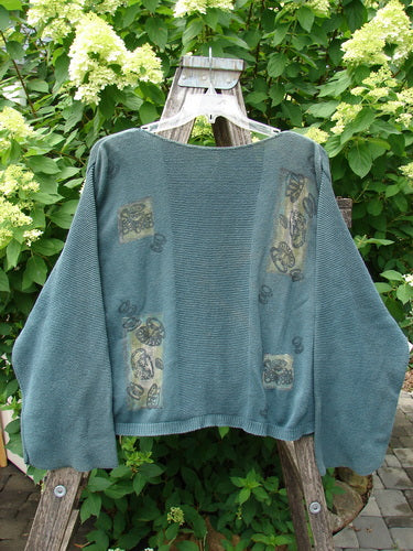 1994 Panel Cardigan Sweater Magic Tea Party Nightshade OSFA features batwing sleeves and decorative patches, displayed on a wooden stand with a lush garden backdrop, embodying vintage Blue Fish Clothing's creative expression.