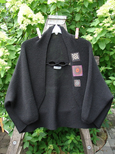 Black Barclay Patched Alpaca Rear Window Shrug OSFA draped over a ladder, featuring a loose, boxy cut, dropped sleeves, folded collar, and colorful patches, held by clothespins against greenery.