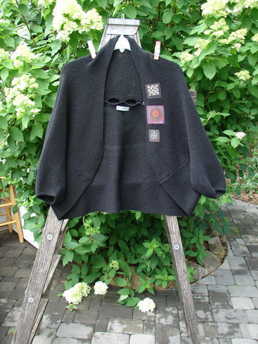 Barclay Patched Alpaca Rear Window Shrug Black OSFA displayed on a wooden ladder, showcasing its loose fit, short kimono sleeves, hood, keyhole neckline, and decorative patches against a garden backdrop.