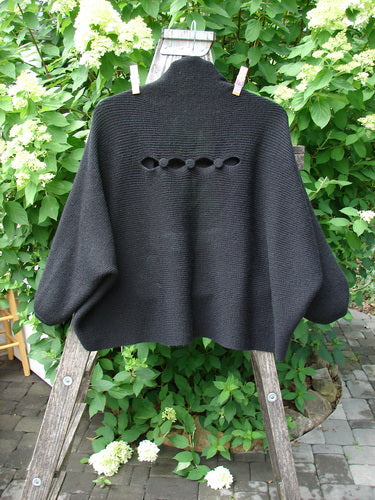 Barclay Patched Alpaca Rear Window Shrug Black OSFA draped on a wooden ladder, showcasing its distinctive eyelet openings and wide sleeves, set against a lush garden backdrop.
