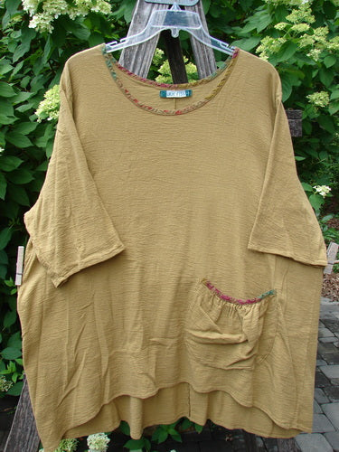 Barclay Gauze Round Bottom Tie Back Pocket Top Ditsy Daisy Golden Size 2, featuring mid-length sleeves, scooped neckline with trim, and a patch pocket, hangs on a plastic hanger against leafy shrubs.