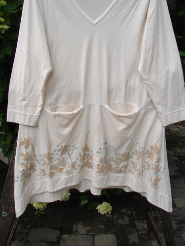Barclay Deep V Neck Rear Banded Pocket Tunic Morning Garden Dew Size 1: A cream tunic with a V-neck and front patch pockets, featuring a floral border print, displayed in a garden setting.