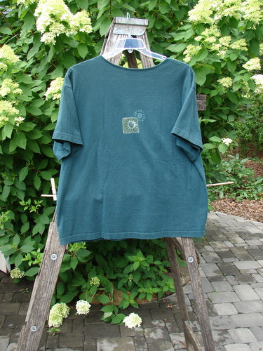 1996 Short Sleeved Promo Tee Old Frenchtown Field Viridian Size 1, featuring a stylized sun and leaf motif, draped over a wooden step-ladder against a lush garden backdrop, highlighting its vintage appeal.