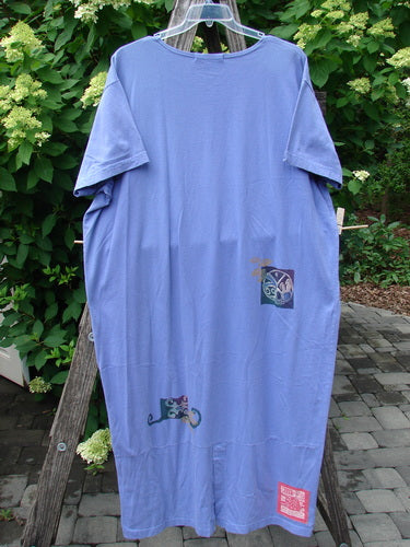 1997 Aquavella Dress Celtic Trio Skylark Size 2 features a loose-fitting, periwinkle-blue design with small graphic patches, displayed outdoors on a hanger amidst a lush garden setting.