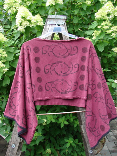 Barclay Cotton Lycra Contrast Crop Shrug features a Celtic Curl design with belled lowers and a laced neckline, showcased on a hanger against a backdrop of lush foliage.