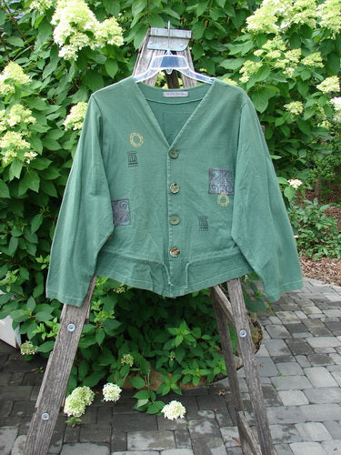 1997 Treehouse Jacket Greek Window Verdigris Size 2: A green cardigan with a double-paneled V-neck, decorative accents, and original buttons, displayed on a ladder against a leafy backdrop.