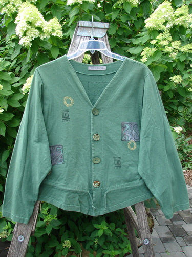1997 Treehouse Jacket Greek Window Verdigris Size 2: A mid-weight organic cotton jacket with a button front, V-neck, unique motifs, and drawcord flounce, hanging on a ladder in a garden setting.