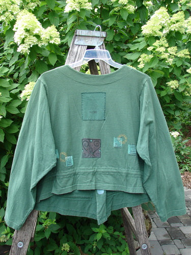 1997 Treehouse Jacket Greek Window Verdigris Size 2, featuring a cropped, forest-green design with decorative patches and a double-paneled V-neck, displayed on a wooden stepladder amidst leafy shrubs.
