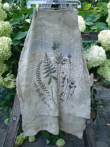 Barclay Crinkle Cotton Scarf Morning Fern Mottled Wheat OSFA, draped on a rustic ladder, showcases fern motifs in a serene garden. The scarf's organic cotton weave adds a touch of vintage elegance.