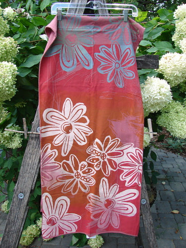 Barclay NWT Wide Sectional Scarf Daisy Desert Rose OSFA, featuring warm hues and daisy motifs, hangs on a wooden trellis, showcasing its organic cotton texture and unfinished rolled edges, perfect for versatile styling.