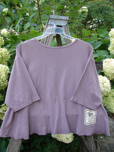 Barclay PMU Patched Short Sleeved Crop A Line Tee Dark Mallow Size 2 displayed on a hanger, showcasing its loose fit, rounded neckline, and unique front alligator-themed patch, set against a lush garden background.