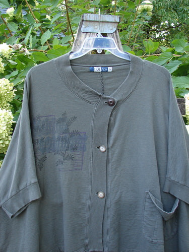 Barclay Slub Dolman Shirttail Pocket Jacket Fern Green Army Size 0, features a loose fit, button-front, dolman sleeves, and a single pocket, hanging against foliage and hydrangeas.