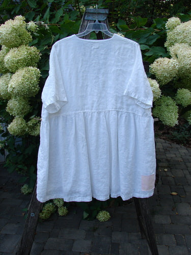1999 Linen Adjustable Wrap Dress Fish White OSFA, loose-fitting and short-sleeved, hangs on a clear hanger in a garden setting, framed by hydrangeas and foliage, accentuating its airy, artistic design.