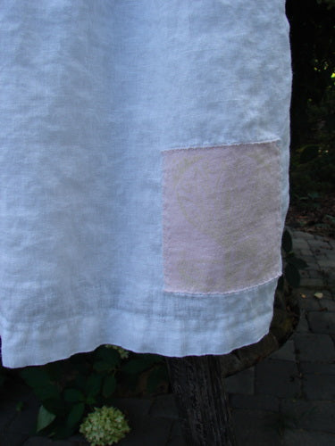 1999 Linen Adjustable Wrap Dress Fish White OSFA draped over a branch, showcasing a delicate pink patch near the hem. This vintage wrap dress features a flowing skirt and pearl button accents.