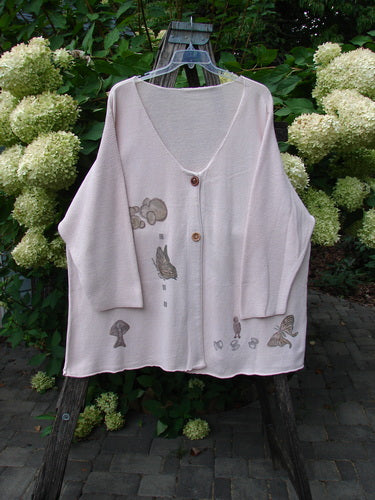 1994 2 Button Cardigan Sweater Magical Garden Pink Granite OSFA draped on a ladder, featuring a deep V-neck with wooden buttons, whimsical prints, and varying stitches, embodying vintage charm amidst foliage.