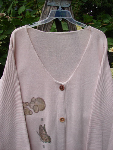 1994 2 Button Cardigan Sweater Magical Garden Pink Granite OSFA, featuring a deep V-neck, two wooden-tone buttons, drop shoulders, and a cloud-like motif with butterfly design, hangs outdoors.