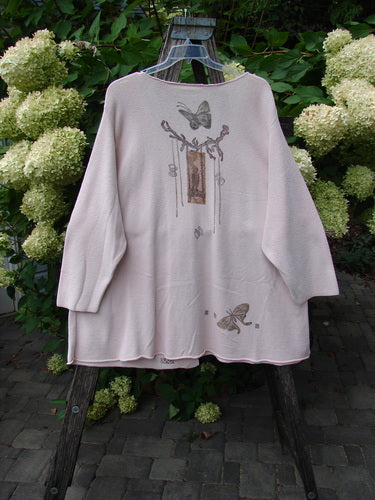 1994 2 Button Cardigan Sweater Magical Garden Pink Granite OSFA displayed on a wooden easel outdoors, showcasing a butterfly and branch motif on the back, amidst lush greenery and hydrangeas.