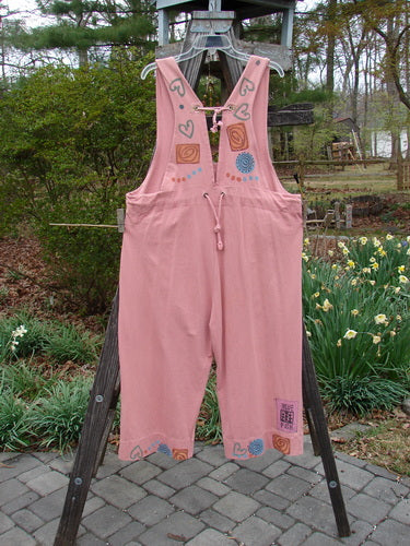 1993 Railroad Overall Pant Jumper Hearts Dried Rose Size 1 hanging on a wooden rack, showcasing its deep oval neckline, front pockets, paneled lowers, and heart and swirl painted design.