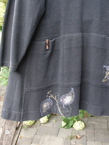 1998 Heavy Berber Kindlewood Tunic Leaf Sprig Black OSFA drapes over a wooden structure, showcasing its subtle earth-toned leaf motif along the hem and a small pocket with a wooden toggle.