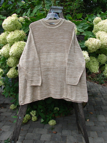 Barclay Jennifer's Rollneck Sweater Marled Aspen OSFA hangs on a wooden display, showcasing its generous A-line shape, rolled hems, and textured knits, set against an outdoor backdrop of foliage and hydrangeas.
