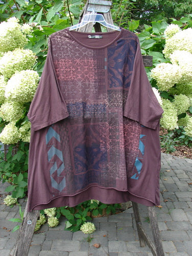 Barclay Art Tee Grid Flower Lower Lily Red Plum OSFA displayed outdoors, showcasing its oversized, tunic-style design with geometric and floral patterns, rounded neckline, and varying hemline against green foliage and white hydrangeas.