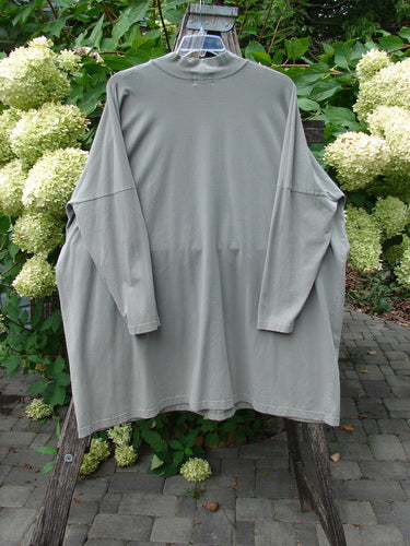 1999 Cotton Lycra Square Mock Pullover Unpainted Grey Stone Size 2 displayed outdoors on a wooden frame, showcasing its loose, boxy shape, dolman drop shoulders, and mock turtleneck amidst green foliage and white blooms.