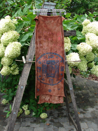 Barclay Diagonal Scarf Wrap Belief Spirit Magic Mottled Rust OSFA draped over a hanger, showcasing its sweet raw curly edges and diagonal seams against lush foliage and hydrangeas.