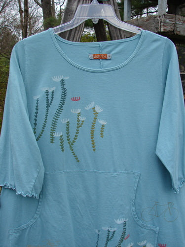 Barclay Three Quarter Sleeved Cafe Pocket Top in Dusty Aqua with white flowers, featuring a feminine neckline, rounded hemline, kangaroo pocket, and three-quarter sleeves.