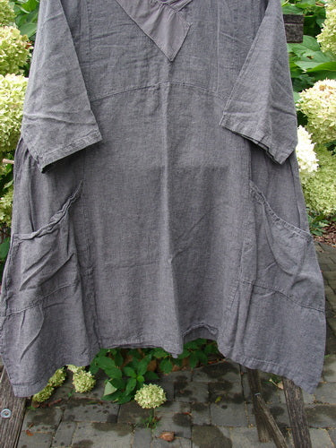 Barclay NWT Linen Cross Over Urchin Dress Stripe Mineral Size 1, featuring a washed-grey fabric, V-neckline, three-quarter sleeves, curved empire waist, and large front pockets, hanging outdoors amid hydrangeas.