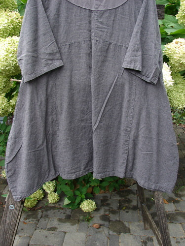 Barclay Linen Cross Over Urchin Pocket Dress Stripe Fig Size 1, displayed on a wooden rack, features a textured grey linen fabric, three-quarter sleeves, a rounded yoke, and an asymmetrical hem.
