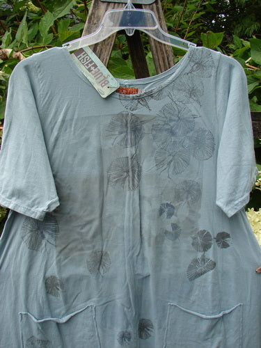 Barclay NWT Seedling Top Dandelion Early Blue Sky Size 1: Loose-fitting pale blue-gray tunic with botanical print, patch pockets, and scalloped hem, hanging against greenery, showcasing Bluefishfinder.com's unique vintage style.