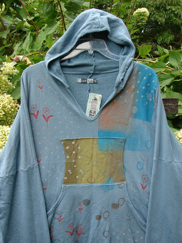 Barclay NWT Fleece Modal Hip To Be Square Hoodie Rain Flower Sea OSFA displayed outdoors, showcasing its V neckline, kangaroo pocket, and unique printed accents against lush green foliage and hydrangeas.