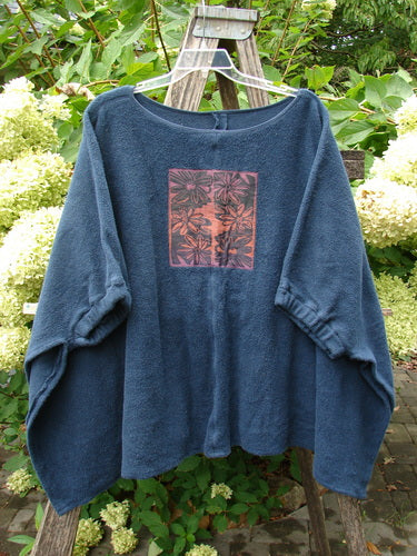 Barclay Celtic Moss Patched Poncho Single Six Daisy Ink Blue OSFA hangs on a wooden ladder, showcasing its loose fit and floral motif square panel. A cozy, versatile pullover with gathered sleeves.
